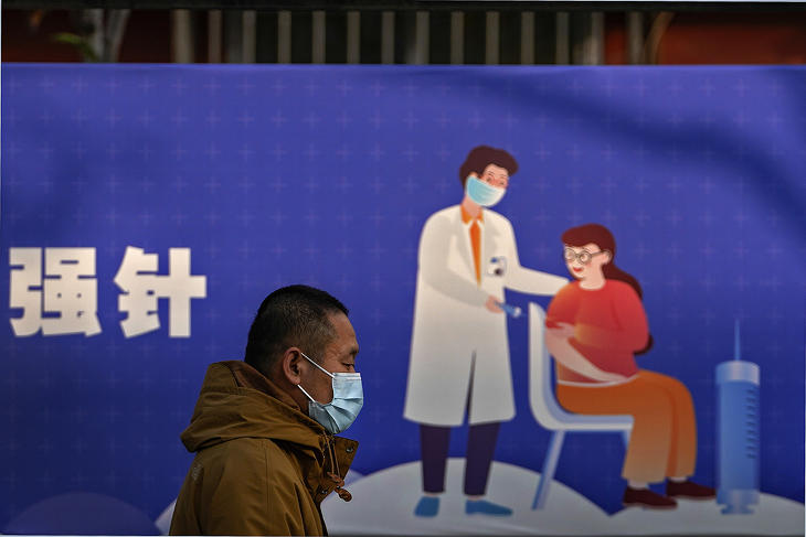 Emlékeztető oltás felvételére biztató óriásplakát előtt halad el egy maszkos férfi – hiába az oltási kampány, messze a nyájimmunitás (Fotó: MTI/AP/Andy Wong)