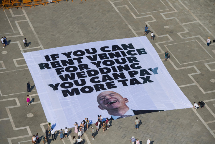 A Greenpeace sajtóirodájának felvételén a nemzetközi környezetvédelmi szervezet aktivistái egy óriási, Jeff Bezos amerikai milliárdos üzletembert ábrázoló transzparenst helyeznek el a velencei Szent Márk téren 2025. június 23-án. A felirat jelentése: „Ha kibérelheted Velencét az esküvődre, adózhatsz is többet”. 