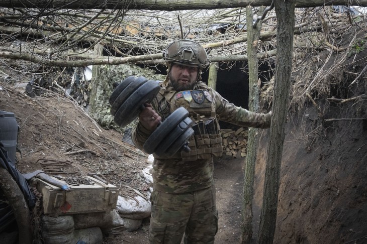 Az ukrán 28. gépesített dandár egyik katonája mutatja súlyzóját lövészárkuk bejárata előtt a kelet-ukrajnai Bahmut település közelében húzódó frontvonalnál 2024. március 3-án. Fotó: MTI/AP/Efrem Lukackij